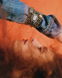 Close-up portrait of a woman wearing layered chain watches and a denim jacket against a warm background.