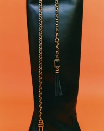 Close-up of a tall black leather boot adorned with a gold chain and tassel belt strap against an orange backdrop