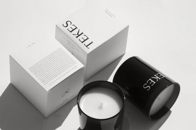 Two Tekes candles and boxes are arranged on a white surface in a minimalist style, evoking a sense of luxury and aromatic design.