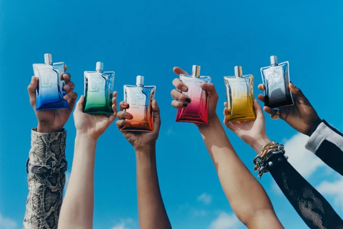 Several diverse arms hold up colorful perfume bottles against a bright blue sky with some clouds, showcasing product diversity and unity.