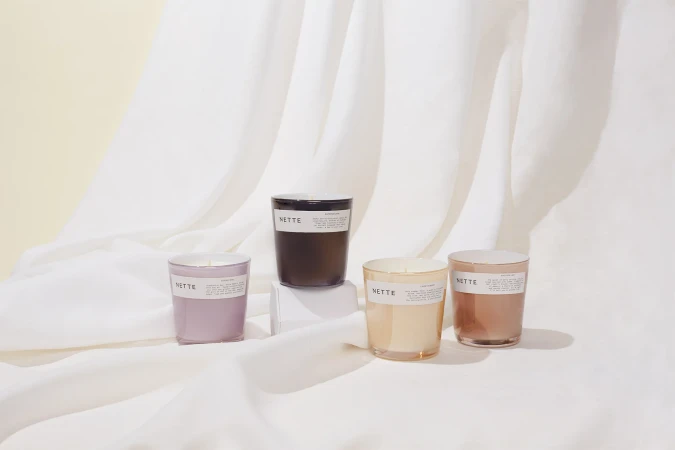 Four translucent glass jar candles with white labels arranged on a white draped fabric surface, set against a light cream background.