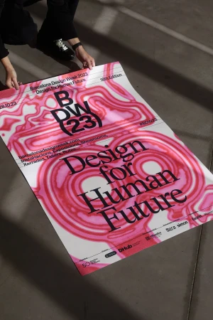 Person holding a pink and white Barcelona Design Week event poster on a concrete floor.