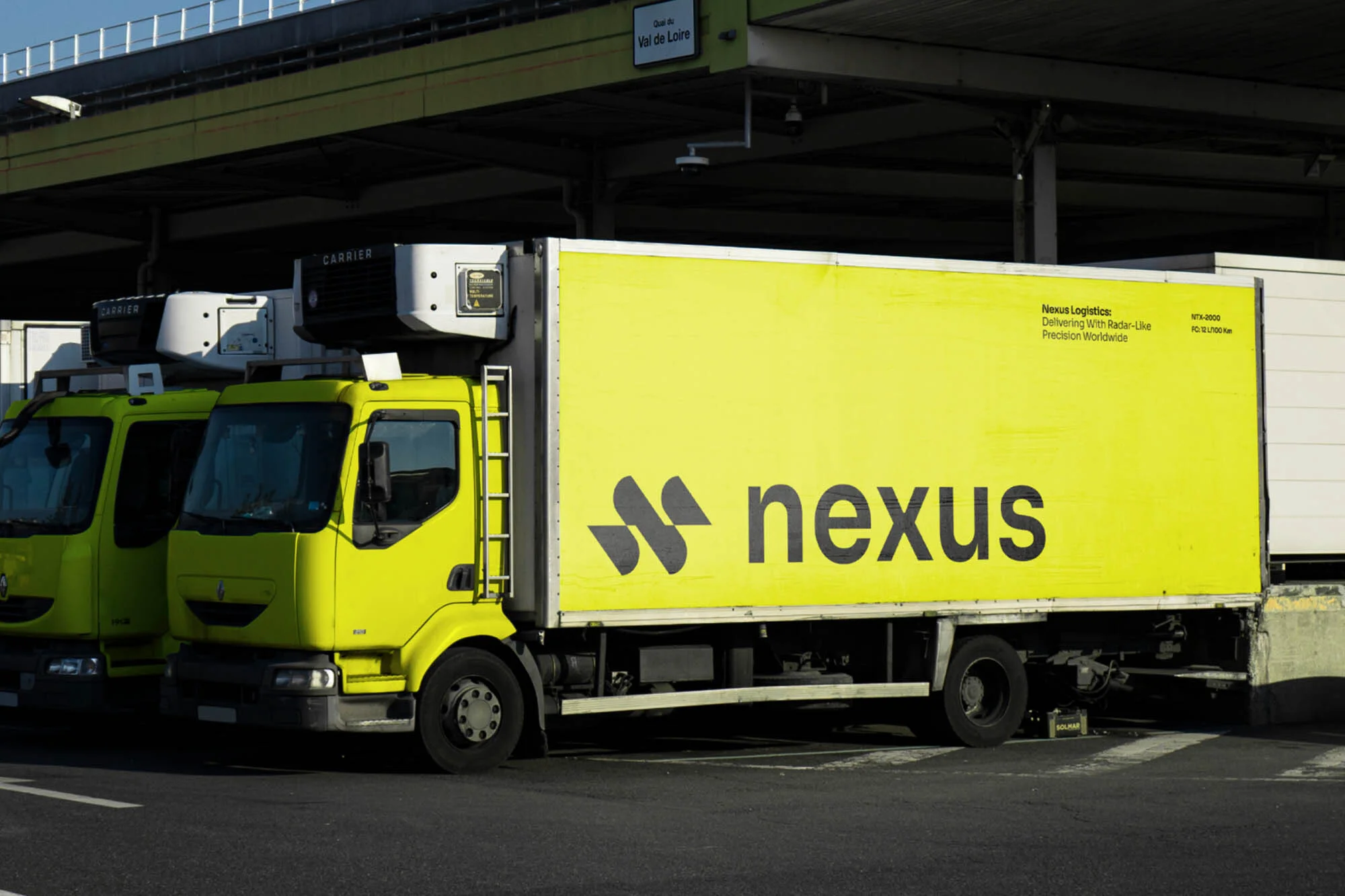 A yellow semi-truck with the word 'nexus' on its trailer parked outdoors on a sunny day. Other semi-trucks are parked nearby.