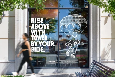 Storefront glass with bold white typography and a line-art parachute graphic, featuring a blurred pedestrian walking past