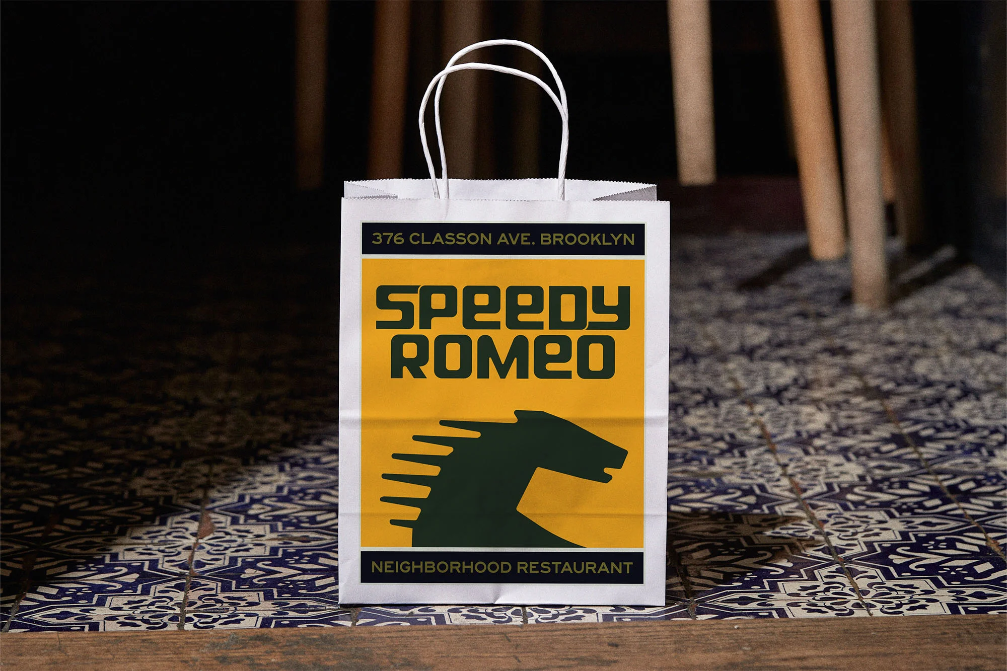 Take-out bag featuring Speedy Romeo restaurant logo with a horse head on a tile surface with wooden chair legs in the background.