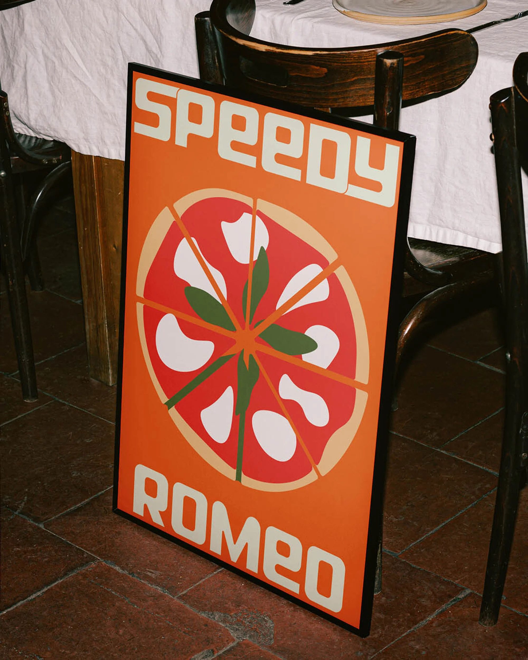 Orange sign on a floor beside a table with chairs features a pizza graphic, and the text “Speedy Romeo” in white sans-serif type.