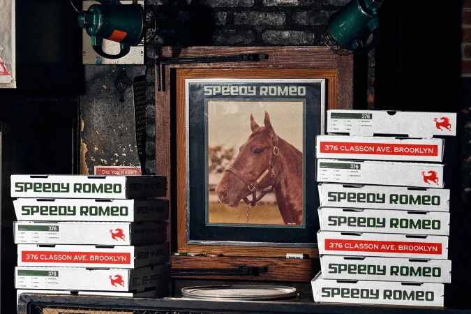 Still life of Speedy Romeo pizza boxes stacked next to a framed picture of a horse in a rustic indoor setting with a brick wall.