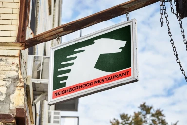 Hanging rectangular restaurant sign with white and green animal logo and red text against a building exterior.