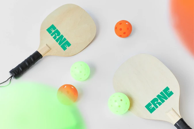 Two pickleball paddles with the word ERNE on them, with two green pickleballs and two orange pickleballs on a white surface.