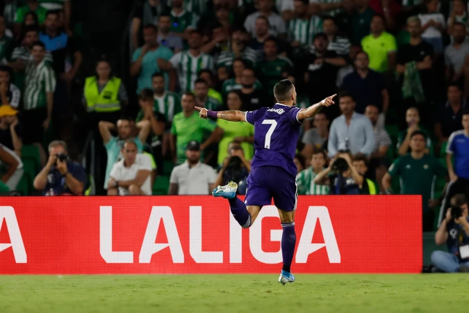 Soccer player celebrates a goal on the field, wearing a purple jersey with the number 7, with a stadium crowd in the background.