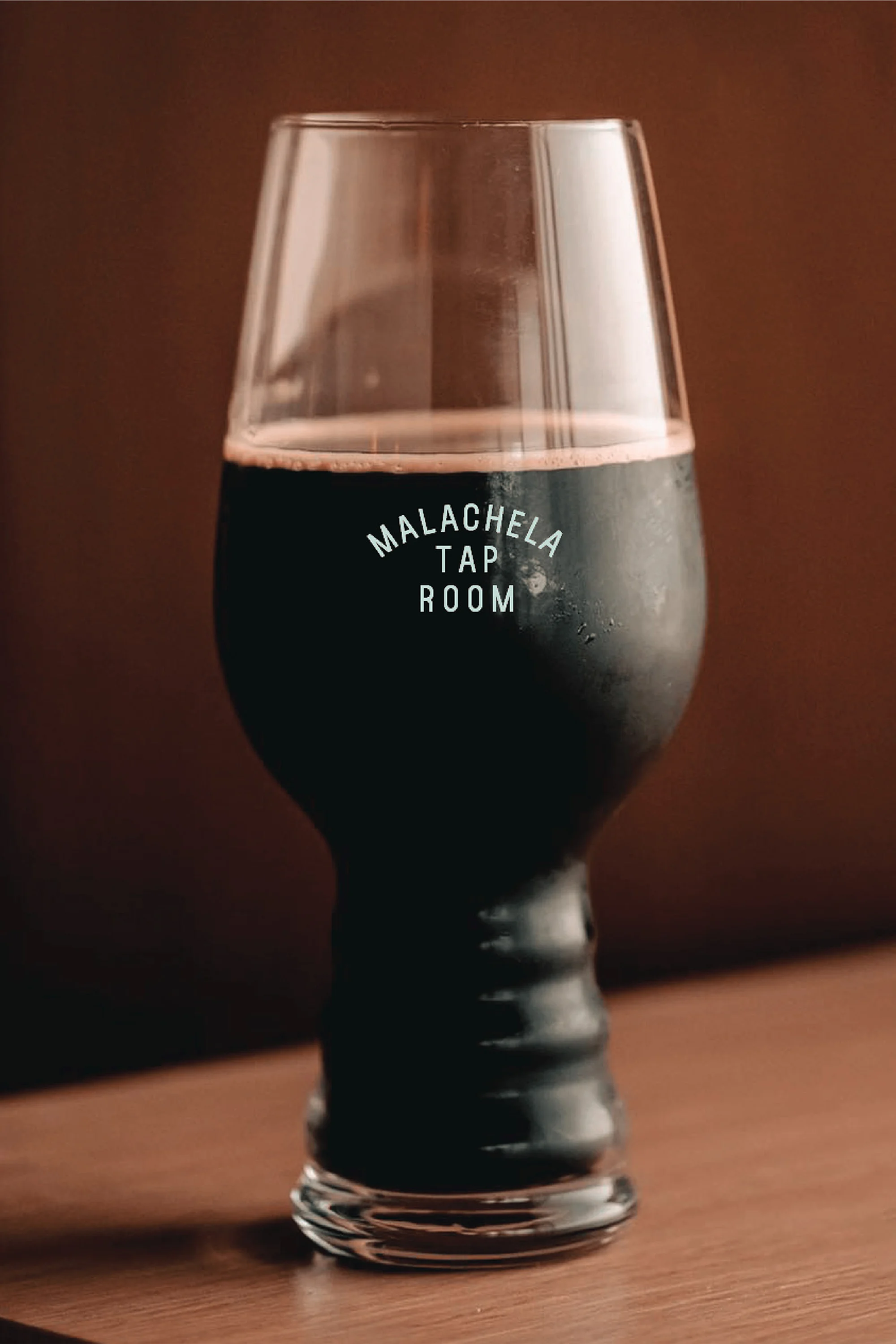 Tulip beer glass filled with dark brown stout, standing on a wooden table in front of a blurred brown background, showcasing beverage.