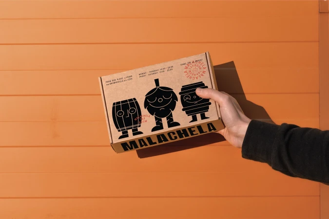 Hand holds Malachela beer box with hop, barrel cartoon art against orange wood. Craft beer concept, brewery marketing, brand visibility.