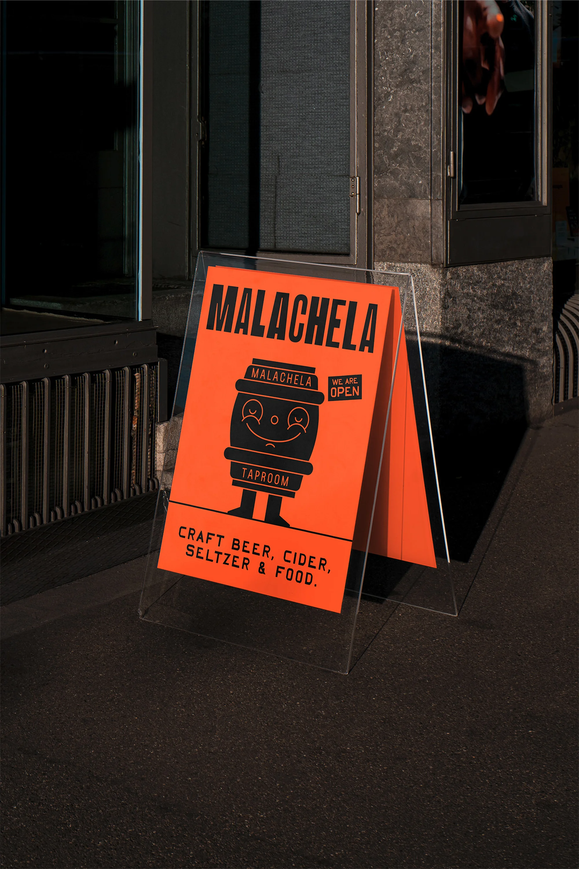 A-frame sign on a city sidewalk advertises Malachela craft beer, cider, seltzer, and food; illustrated with a smiling beer barrel character.