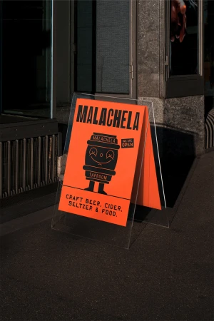 A-frame sign on a city sidewalk advertises Malachela craft beer, cider, seltzer, and food; illustrated with a smiling beer barrel character.