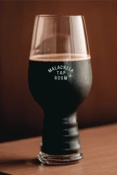 A glass of dark stout beer with foam resting on a wooden surface against a warm brown background.