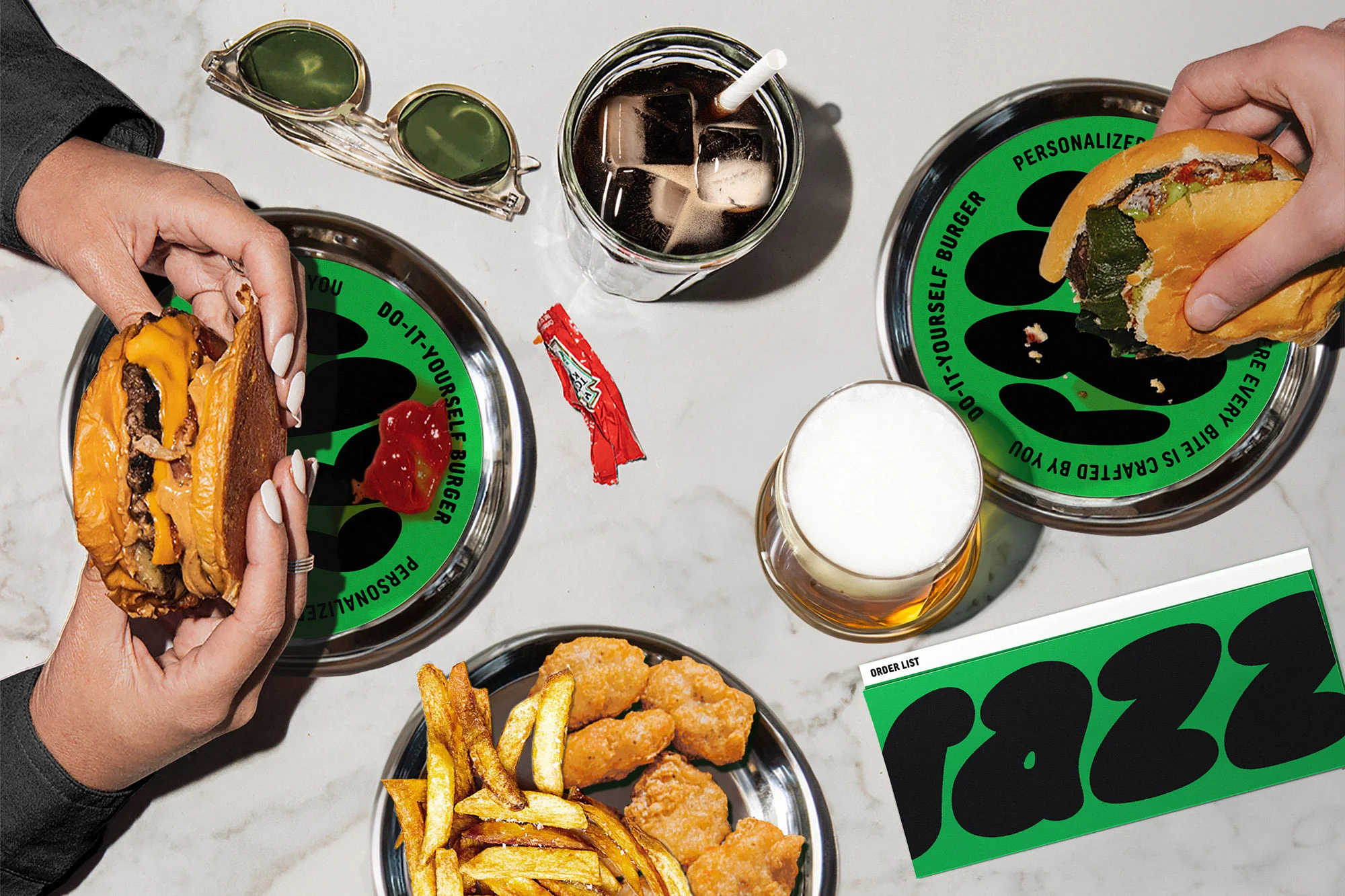 Overhead shot of two people eating burgers and fries on a marble table with drinks, sunglasses, and a menu for 'Razz' restaurant.