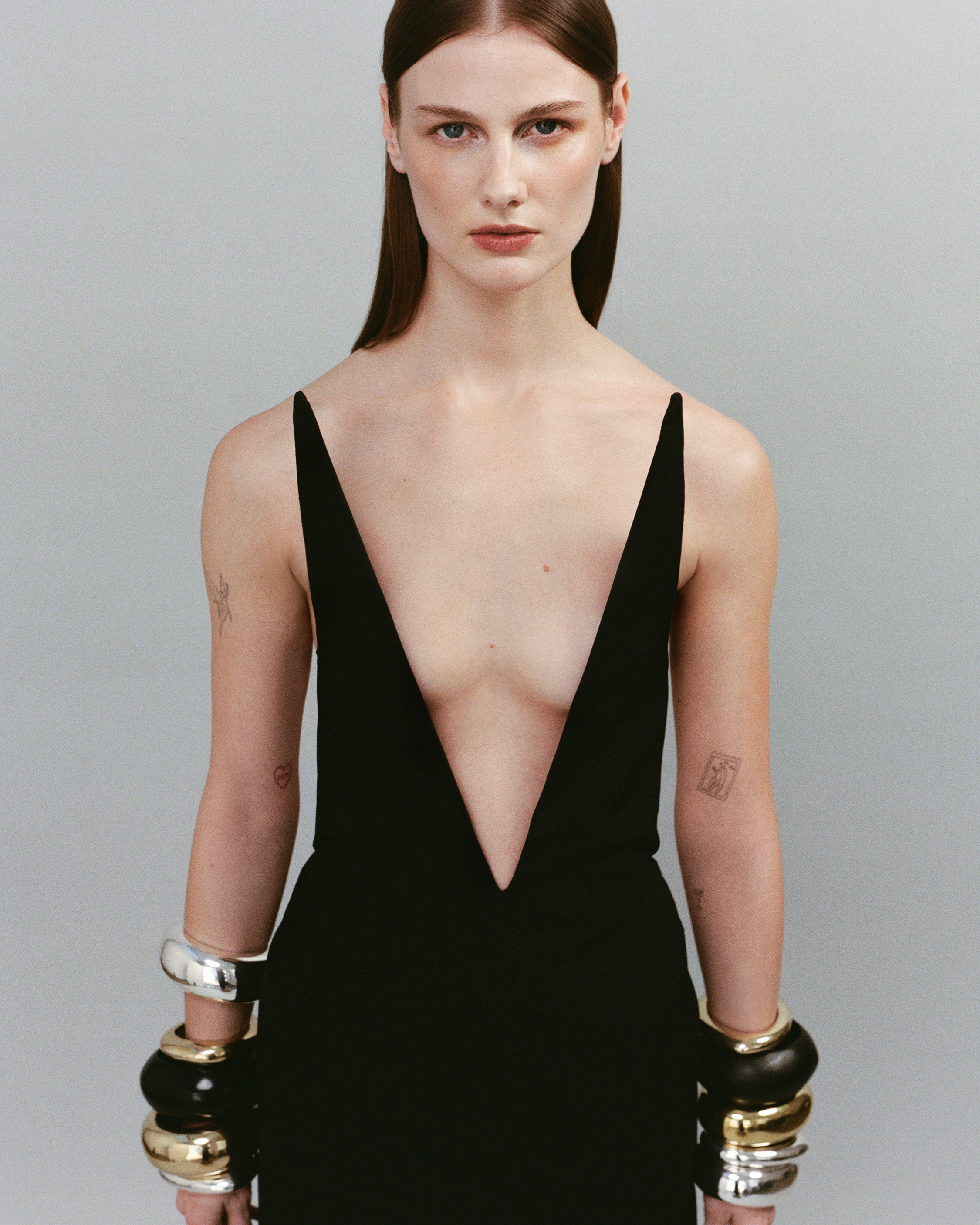 A striking fashion shot of a woman in a black dress and chunky metal bangles set against a neutral backdrop in portrait orientation.