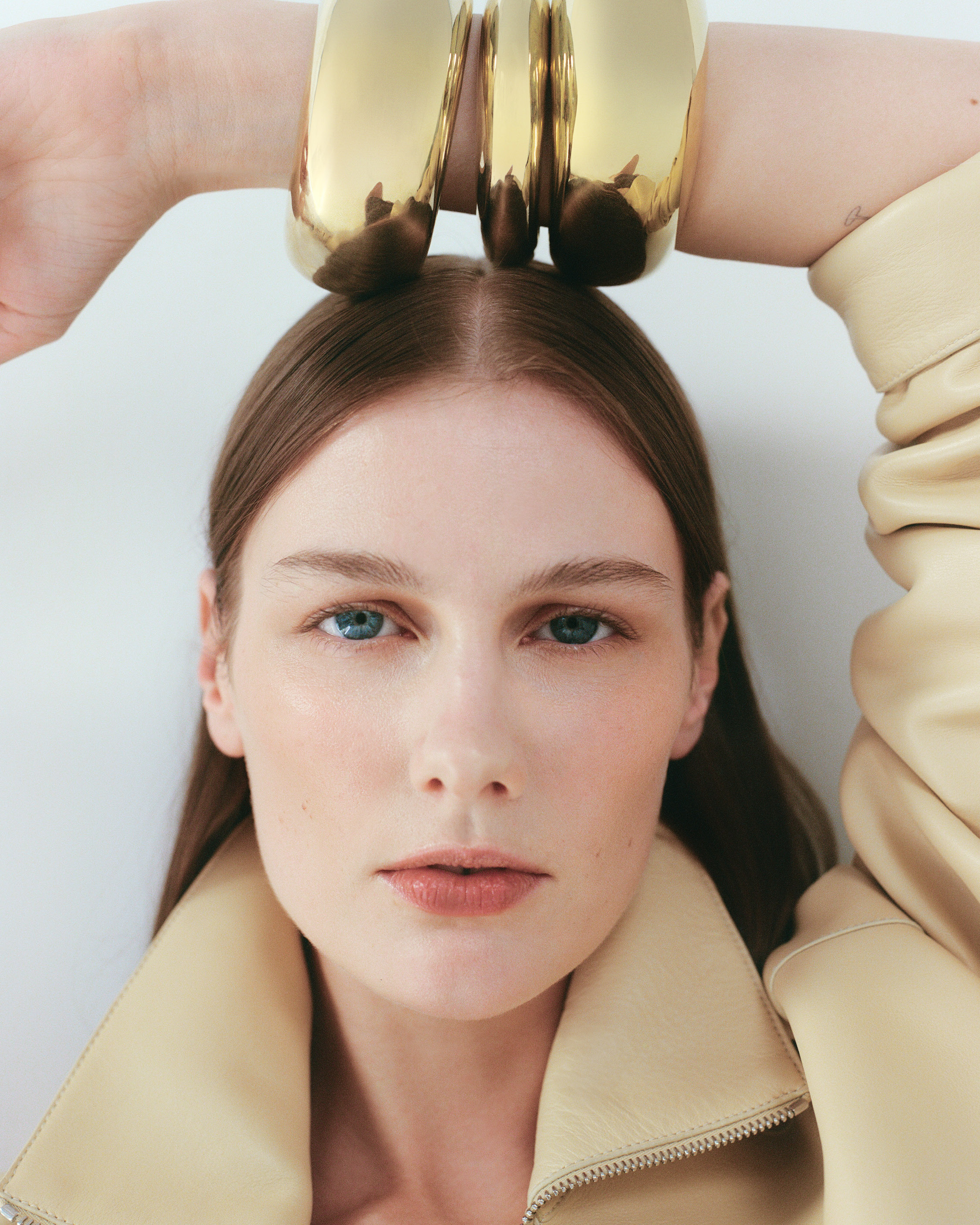 Close-up portrait of a woman with blue eyes wearing a beige jacket, with large gold bangles over her head against a white background.