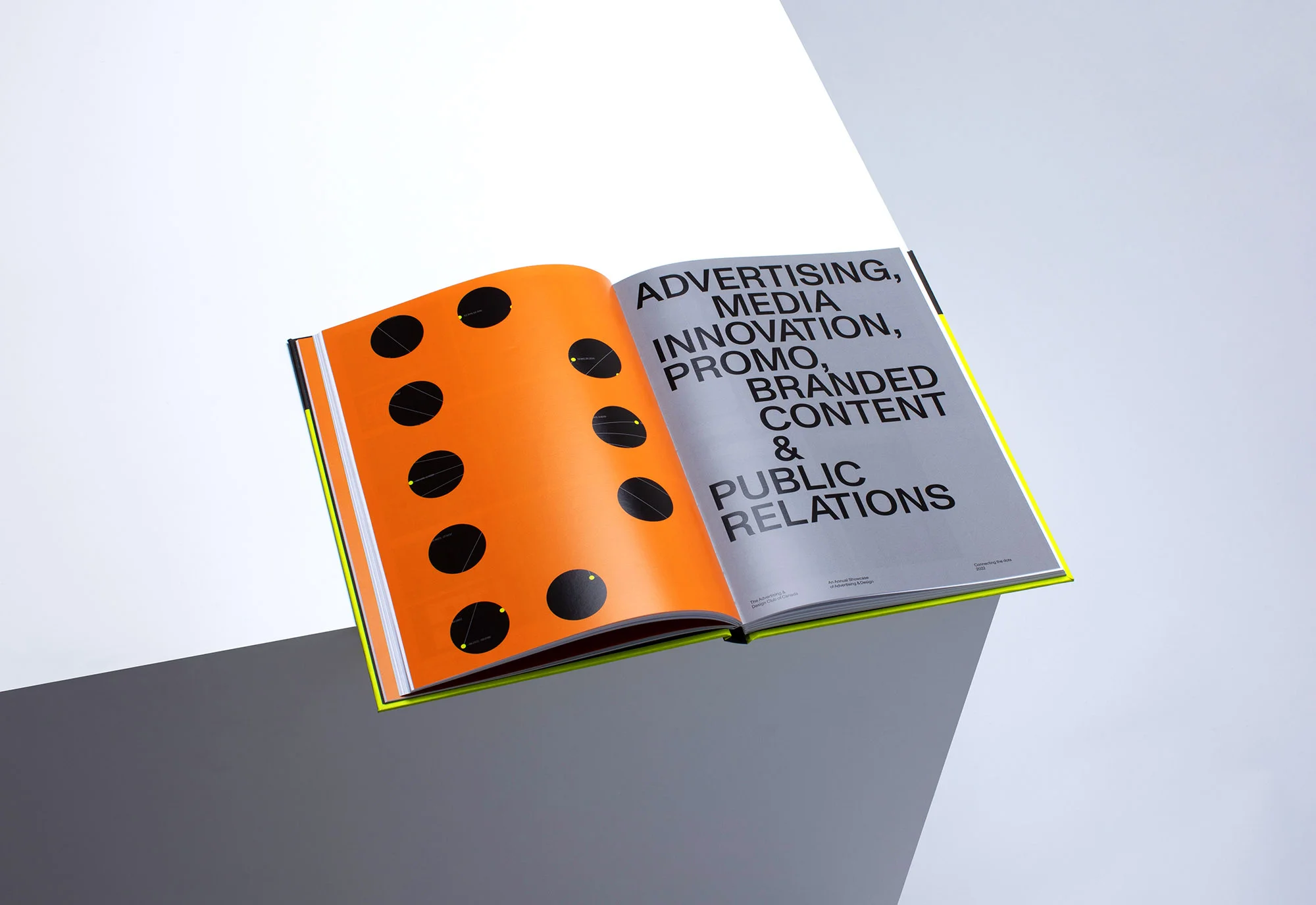 Open book with orange page featuring black circles, contrasting with a grey page filled with black advertising and media text.