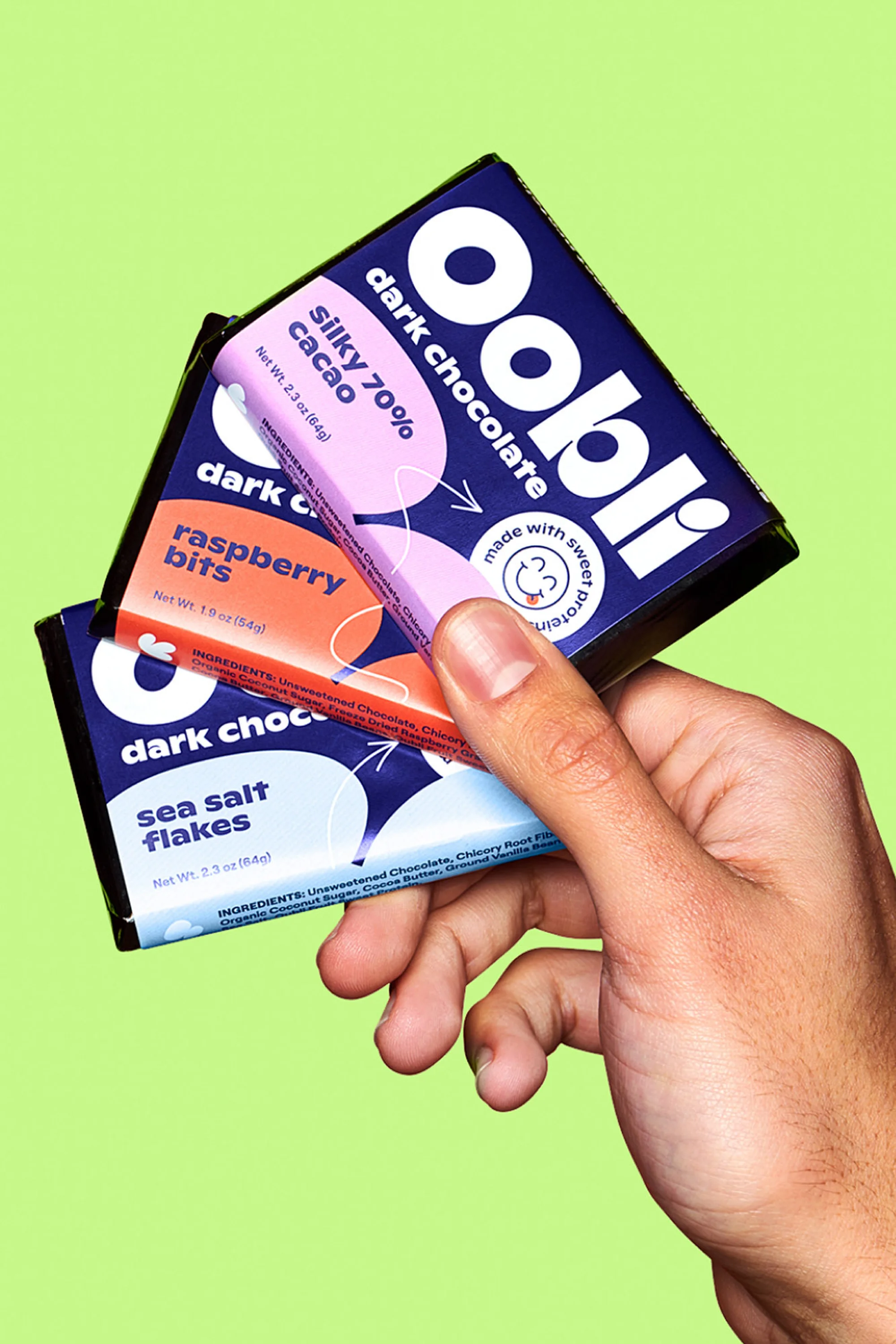 Hand holding three bars of Oobli brand dark chocolate: silky 70% cacao, raspberry bits, and sea salt flakes against a green background.
