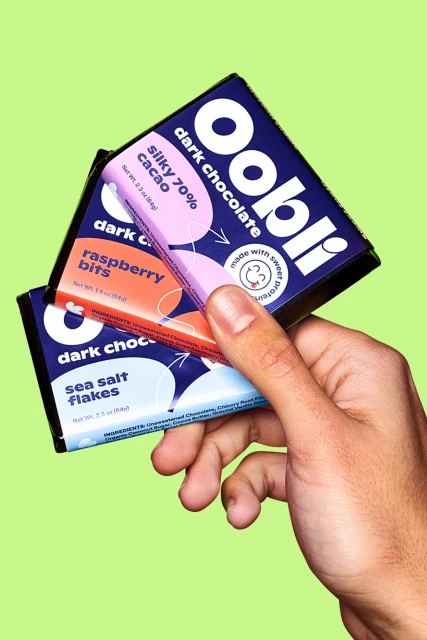 Hand holding three bars of Oobli brand dark chocolate: silky 70% cacao, raspberry bits, and sea salt flakes against a green background.