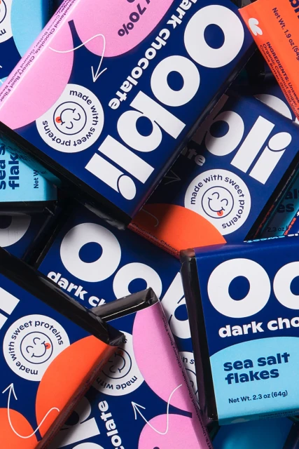 Close-up of Oobli dark chocolate bars in blue and pink packaging, highlighting the brand and product flavors like sea salt flakes.