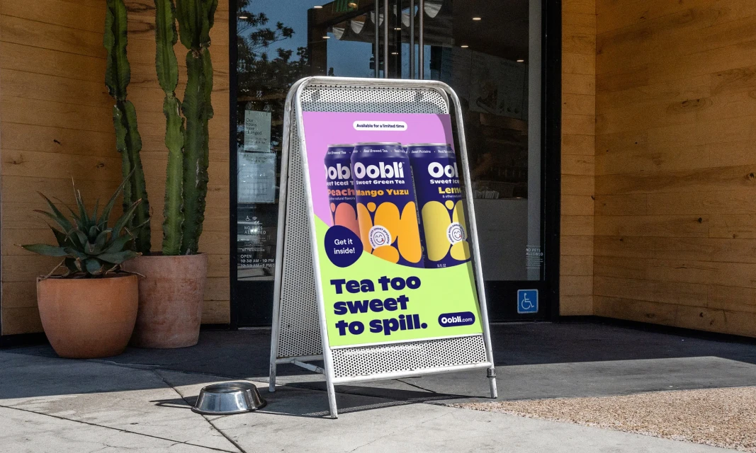 Oobli iced sweet green tea poster on an A-frame sign in front of a building with potted plants. Peach, mango, and yuzu flavors.