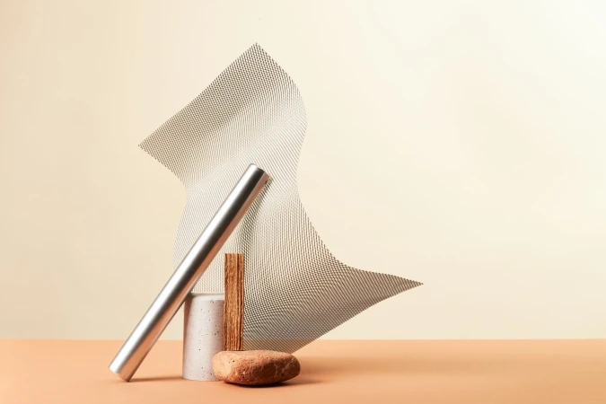 Architectural still life sculpture featuring a curved metal mesh sheet, a silver cylinder, a stone block, wood and a pebble on a beige surface.