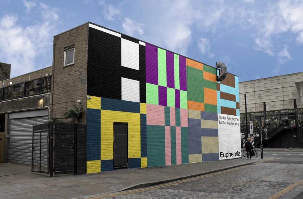 Colorful geometric mural on a brick building in an urban setting. Pedestrians walk by on the sidewalk near the painted artwork.