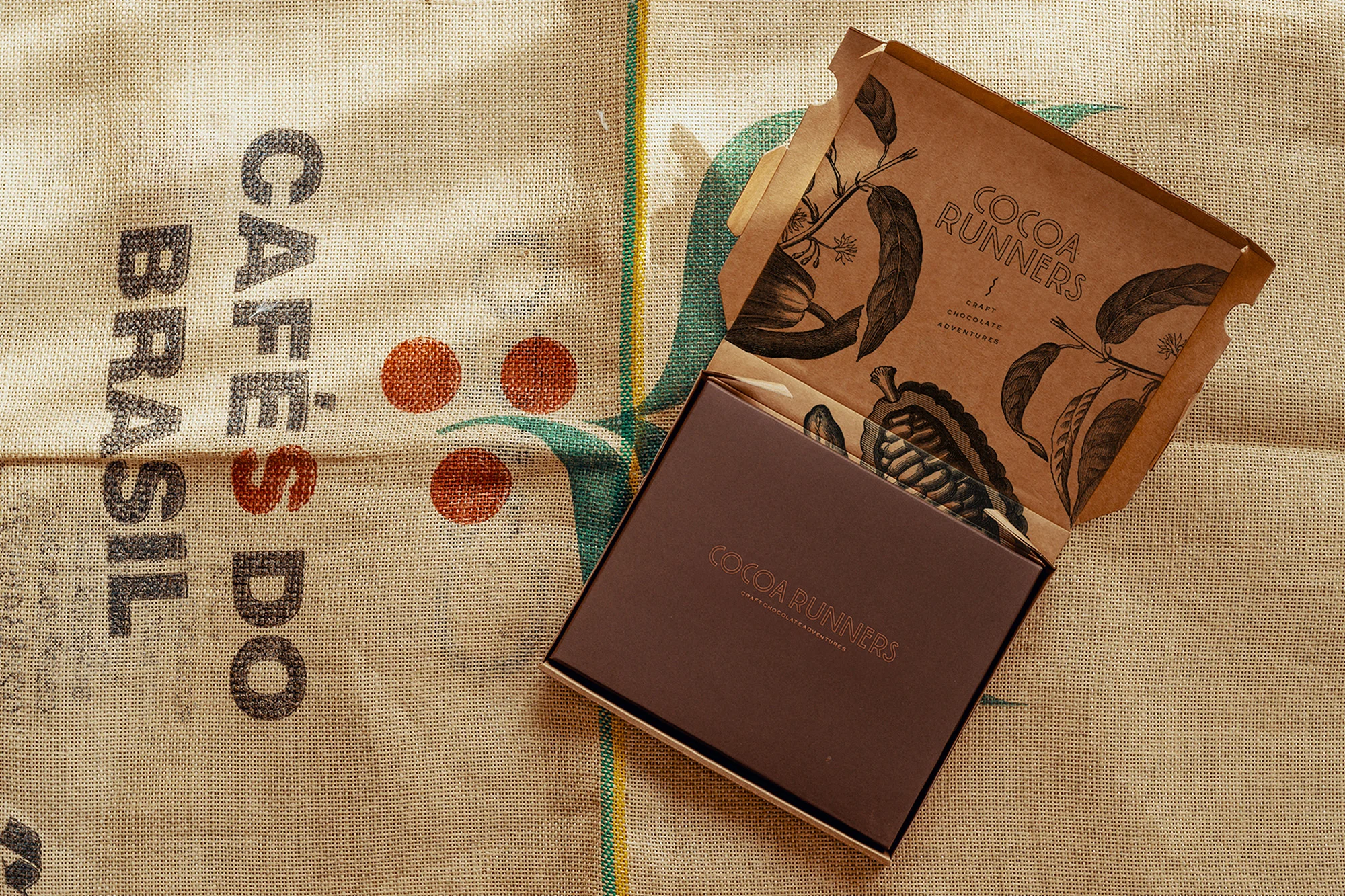 An open box of Cocoa Runners craft chocolate sits on a burlap sack with coffee bean graphics and text 'Cafes Do Brasil'.