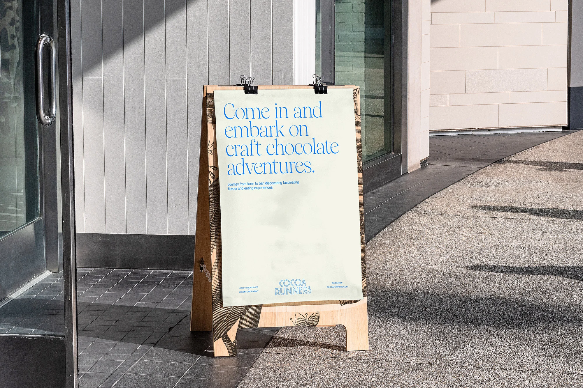 An advertising board for Cocoa Runners stands on a sidewalk next to a building, promoting craft chocolate adventures and tasting experiences.