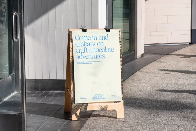An advertising board for Cocoa Runners stands on a sidewalk next to a building, promoting craft chocolate adventures and tasting experiences.