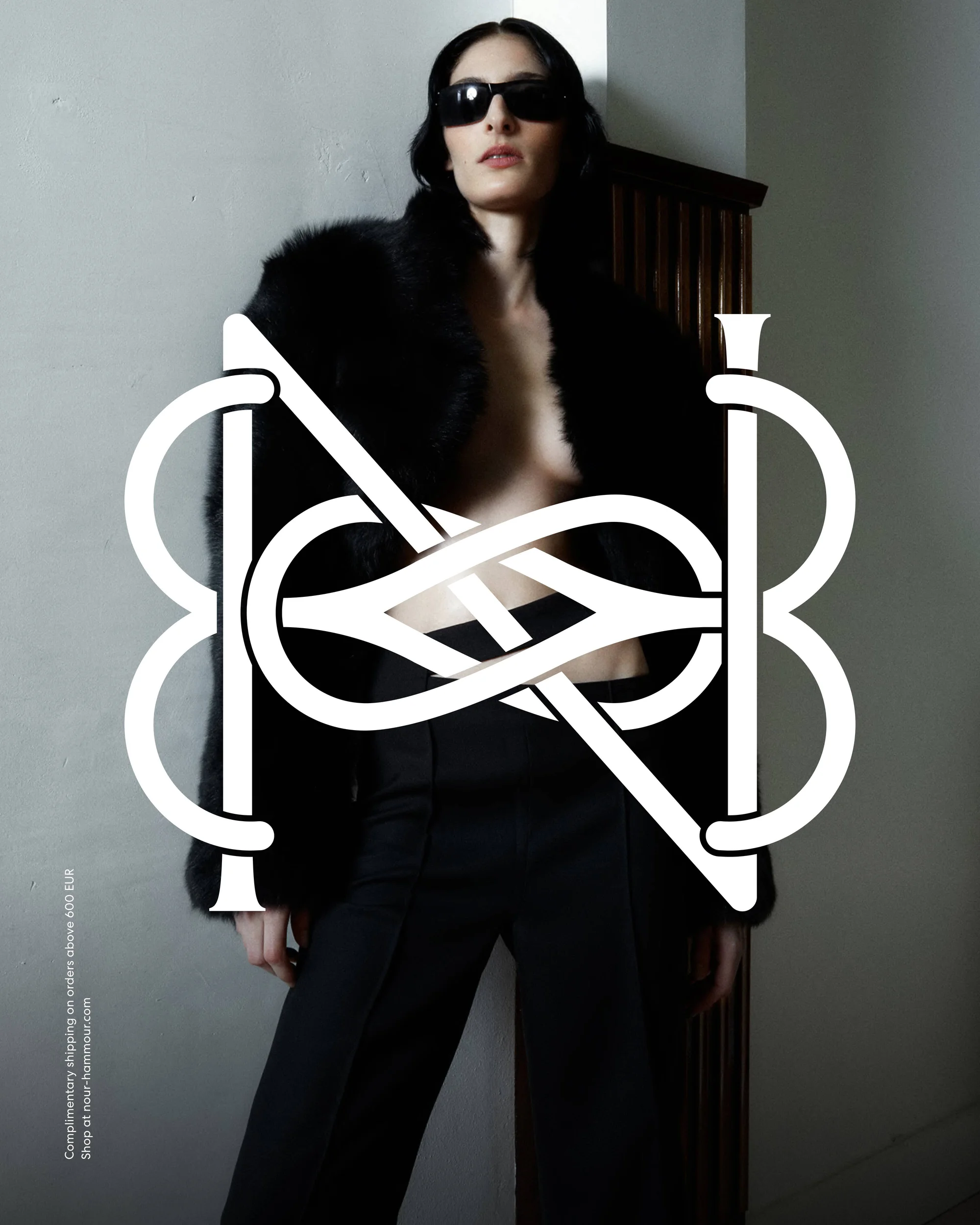 Fashion shot of a woman in a black fur coat and sunglasses, standing in front of a wall with a large, ornate, monogram-like graphic.