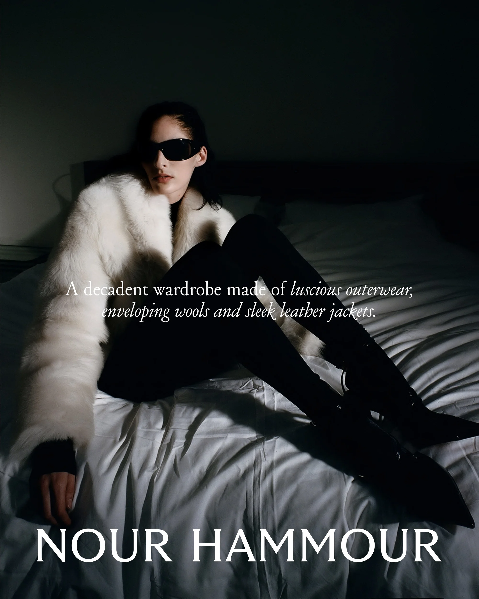 A woman in sunglasses and a white fur coat sits on a bed in a fashion-style portrait. The image features dark boots and leggings.