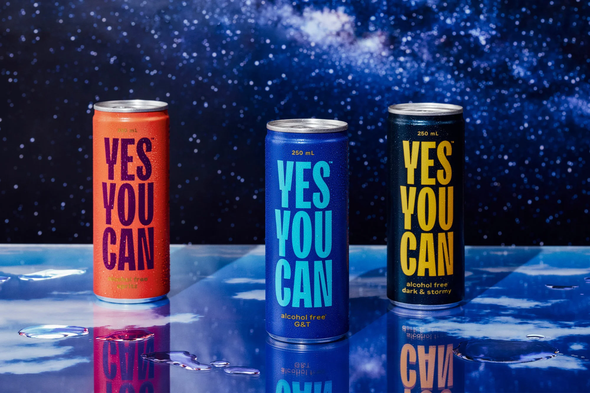 Three colorful 'Yes You Can' beverage cans in orange, blue, and black, placed on a reflective surface against a celestial backdrop.