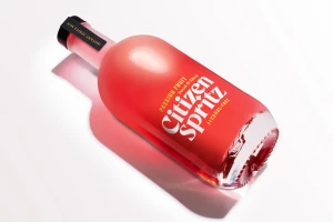 A red glass bottle of Citizen Spritz resting on a smooth white surface, casting a soft shadow with bright studio lighting.