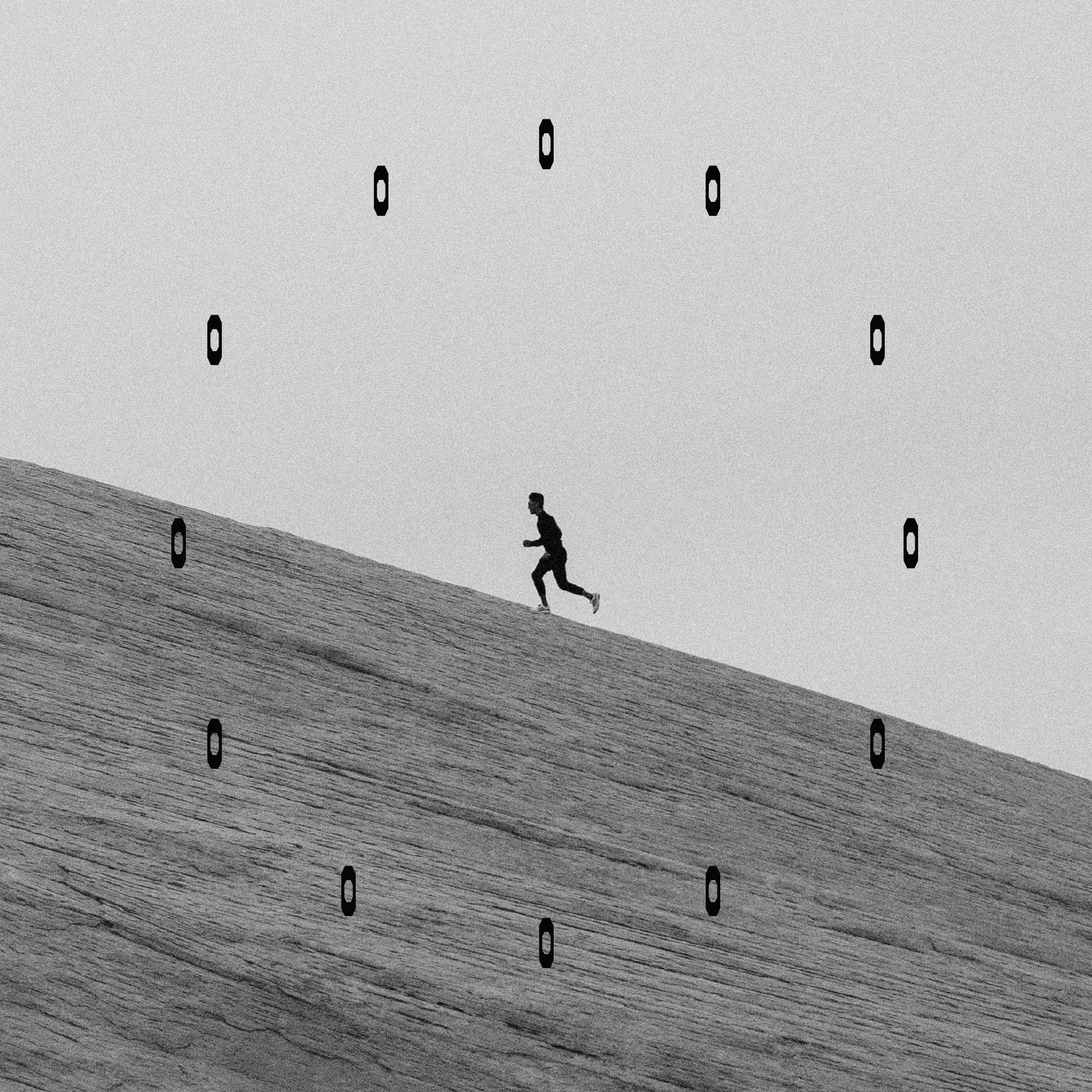 Monochrome surrealist image of a man running up a sand dune with rectangular shapes arranged in a circular pattern in the sky.