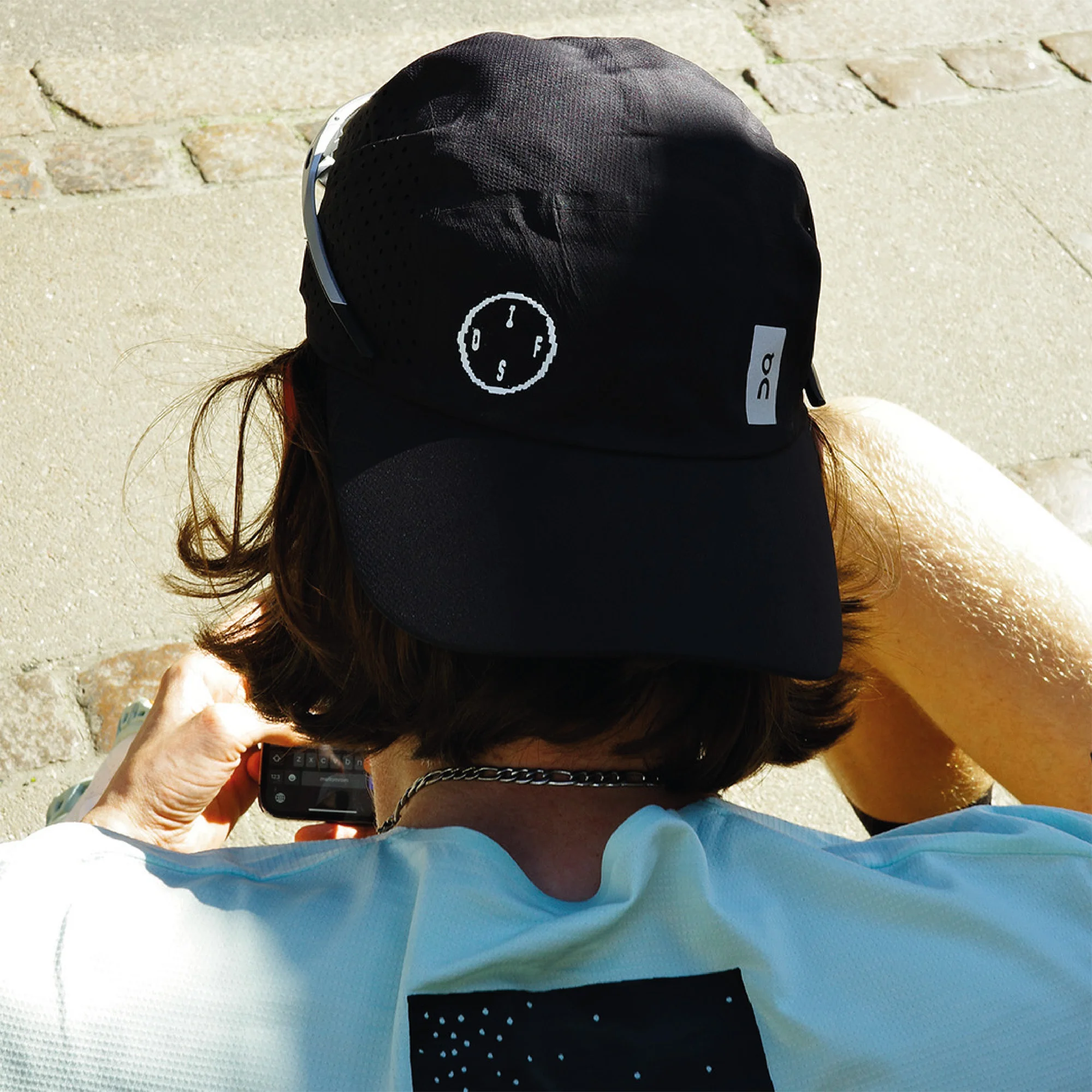 Person with a black cap and a light blue shirt holding a phone, outdoors on a sunny day. The photo is taken from behind.