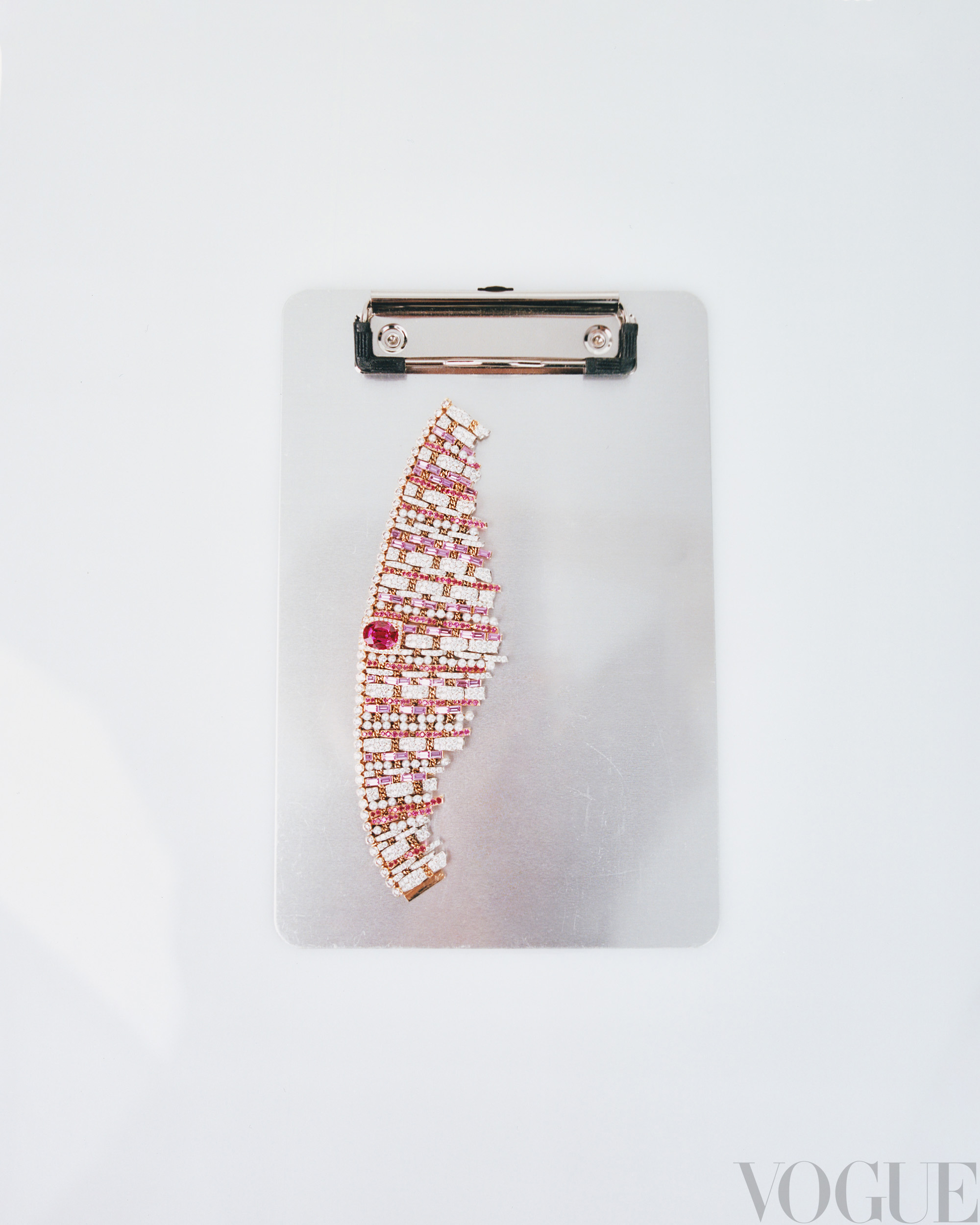 Overhead shot of a clipboard with a semi-circular arrangement of white, pink, and red gem-encrusted jewelry, Vogue logo in lower right.