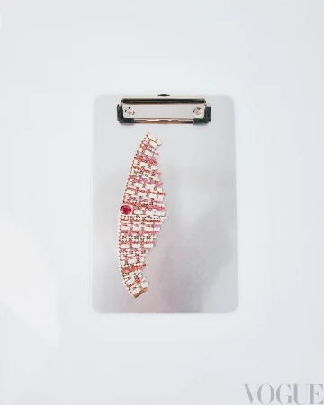 Overhead shot of a clipboard with a semi-circular arrangement of white, pink, and red gem-encrusted jewelry, Vogue logo in lower right.