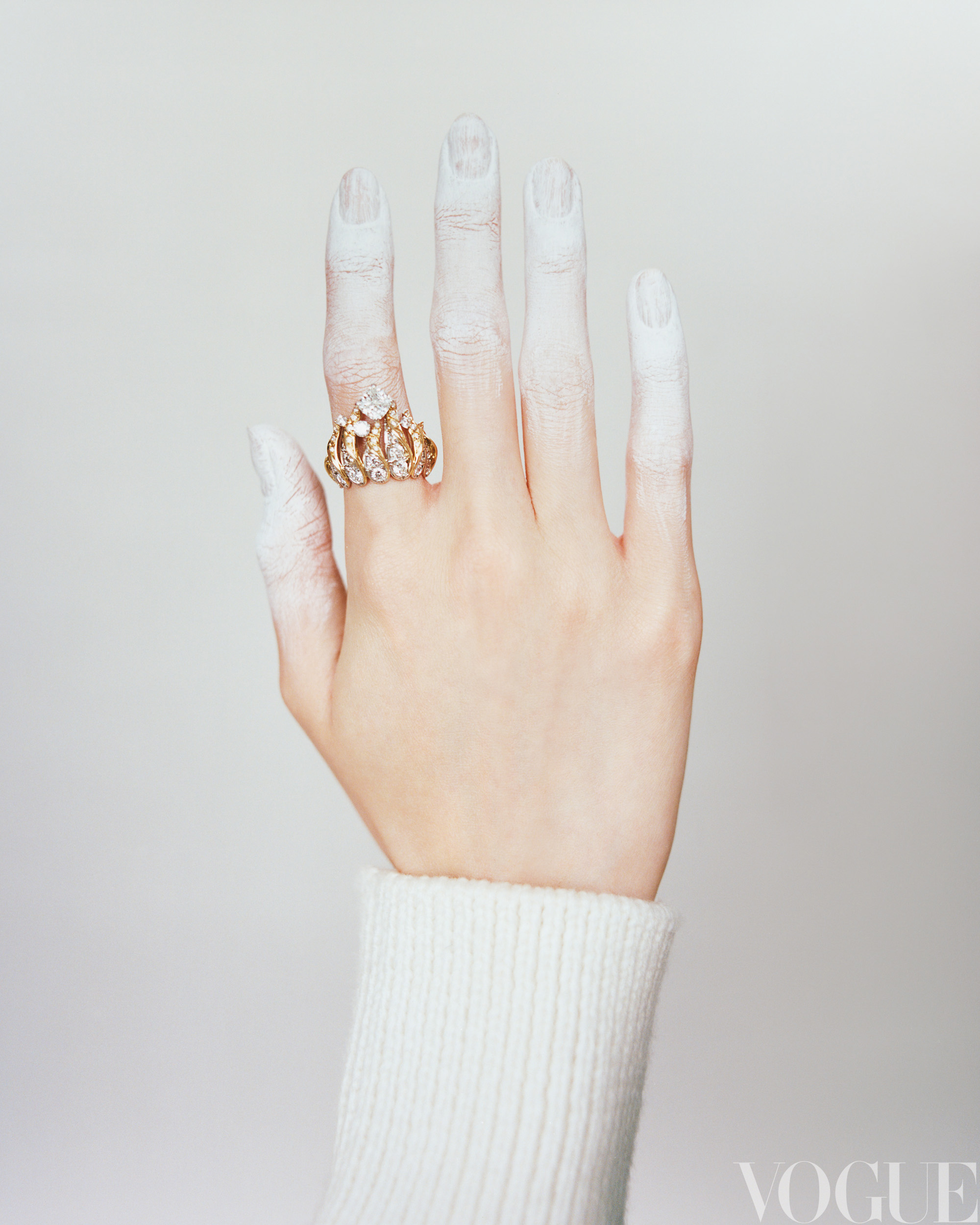 Hand with white paint on fingers wears a gold and jeweled ring against a white background with logo in the lower right corner.