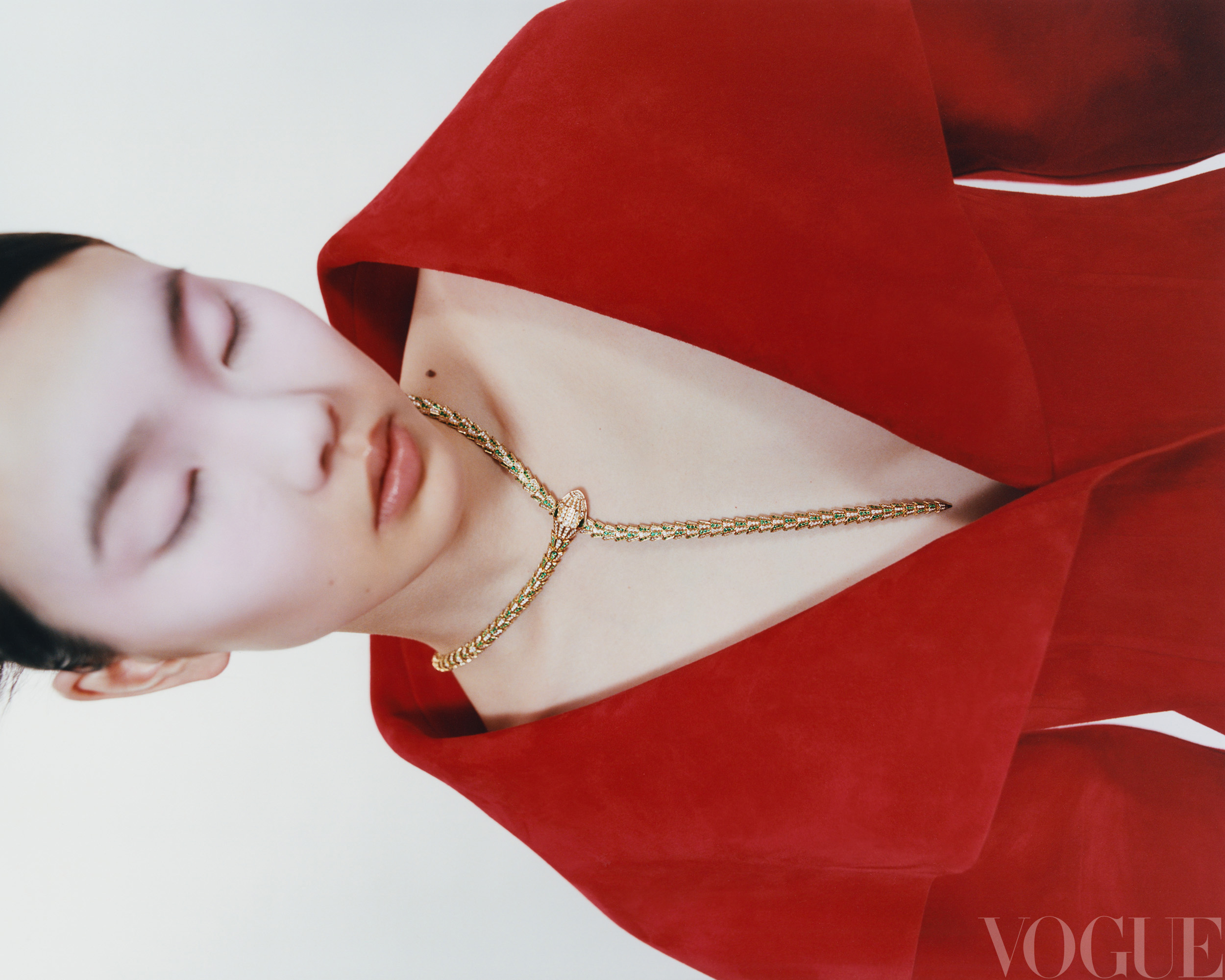 Portrait of an Asian woman with closed eyes wearing a red dress and gold necklace with a pink stone against a white backdrop.