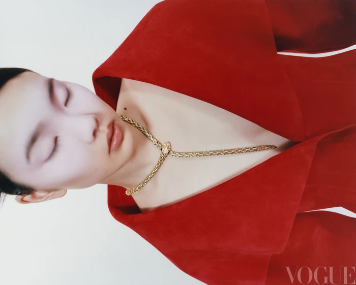 Close-up portrait of a model with closed eyes wearing a bold red jacket and a detailed gold snake necklace against a white background.