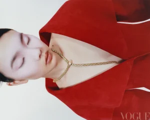 Close-up portrait of a model with closed eyes wearing a bold red jacket and a detailed gold snake necklace against a white background.