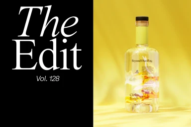 A glass liquor bottle labelled Citrus Bomb by Beyond Distilling stands on a yellow surface against a yellow background beside a black panel with white serif text reading The Edit Vol. 128.