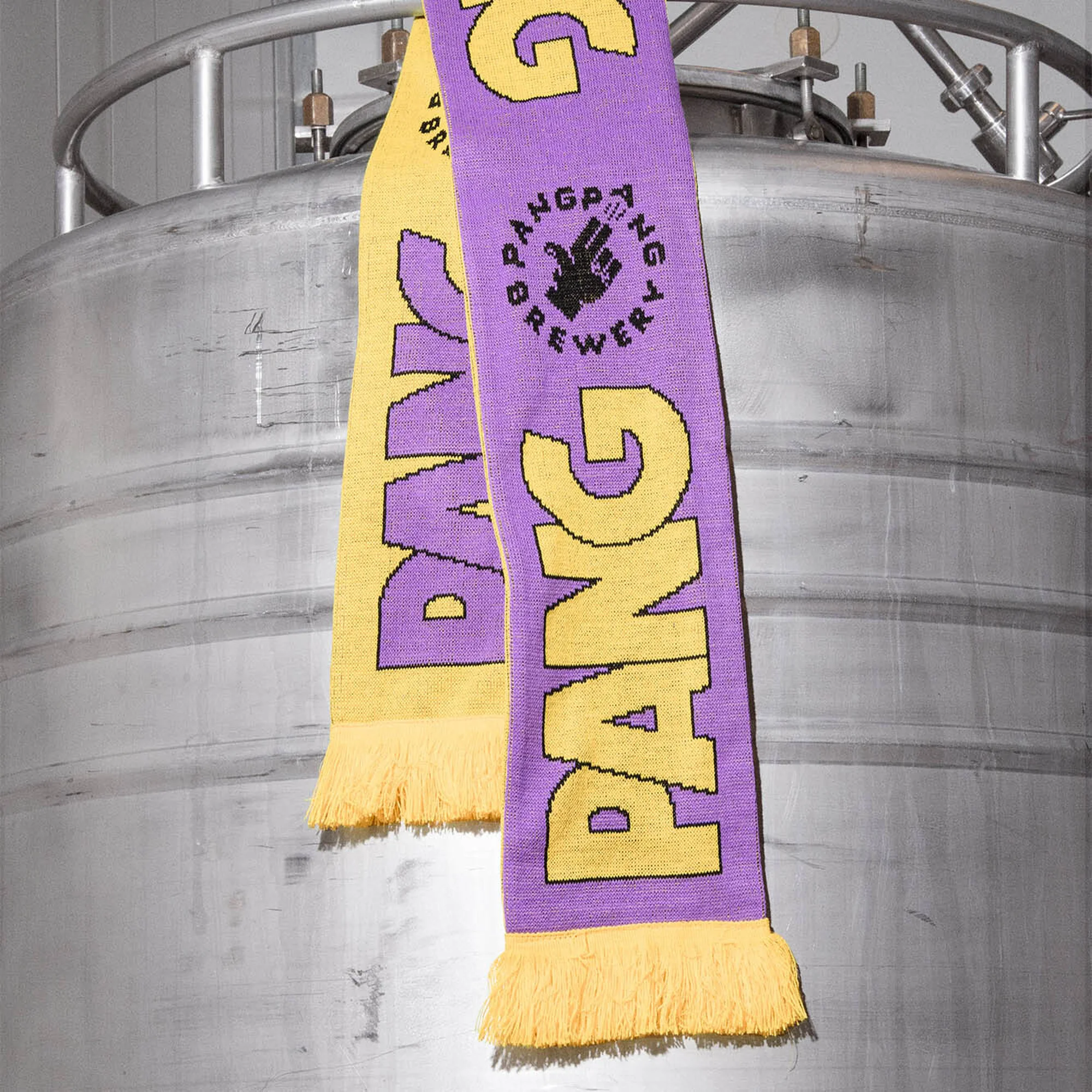 Two PangPang Brewery scarves in yellow and purple with fringe hanging against a large steel brewing tank inside the brewery.