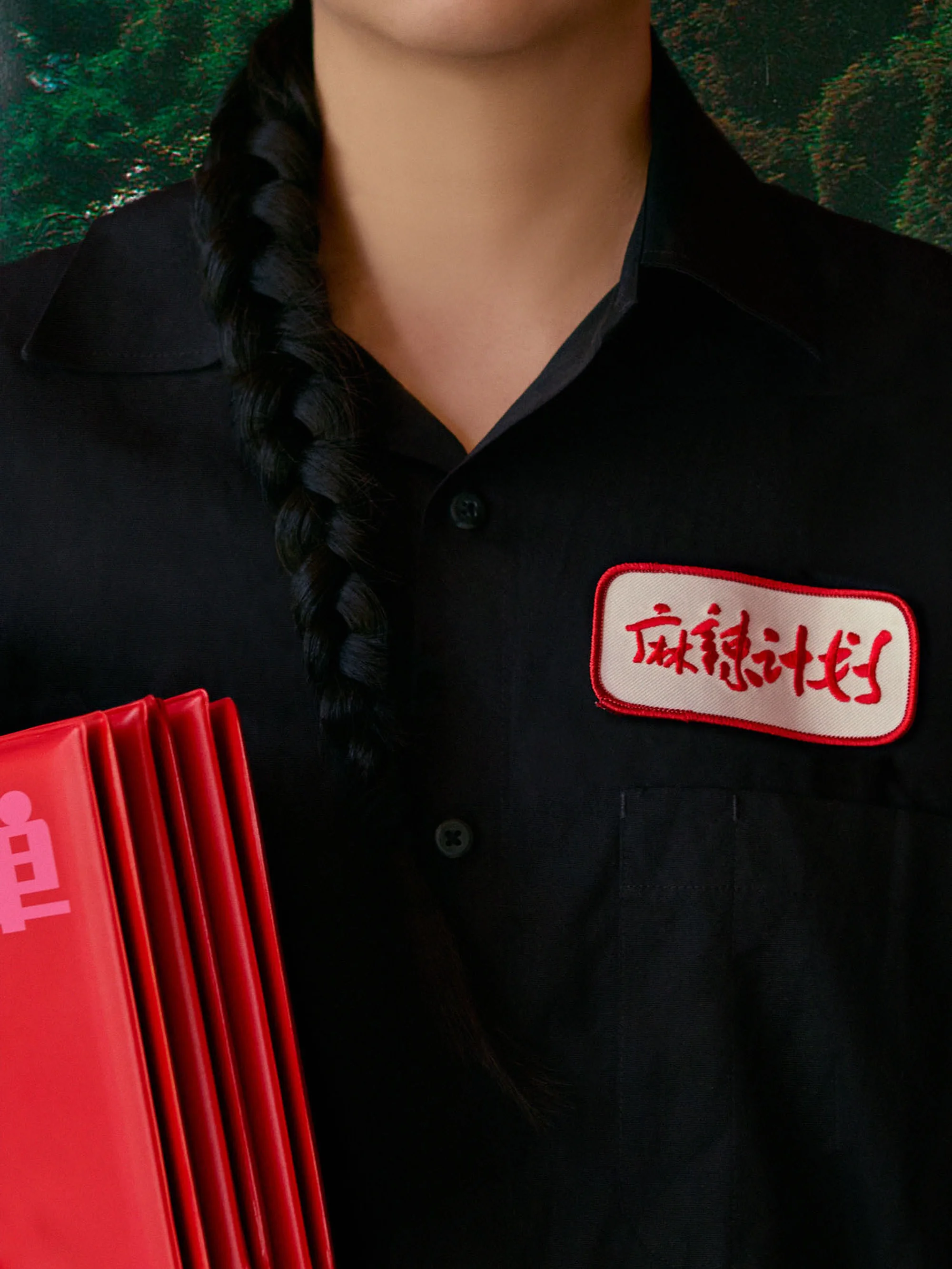 Close-up shot of a person in a black shirt with a braided hairstyle, holding red objects against a leafy green backdrop.