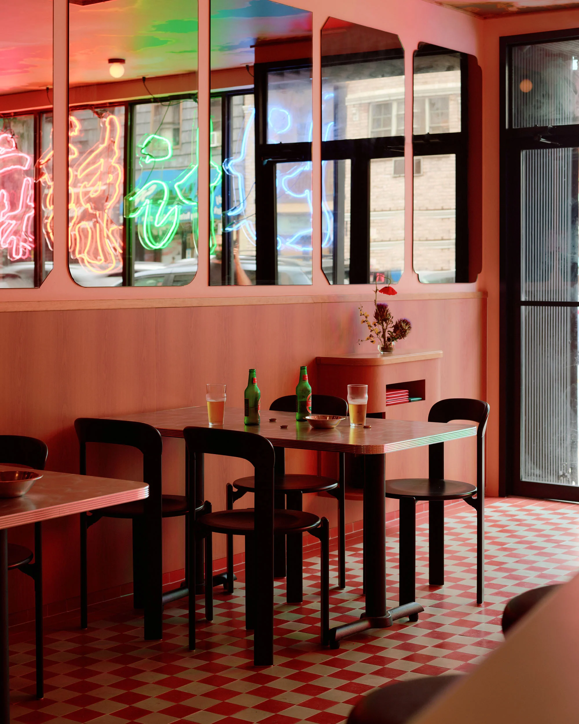 Interior view of a restaurant dining area with tables, chairs, a pink color scheme, and neon signs reflected in mirrors.