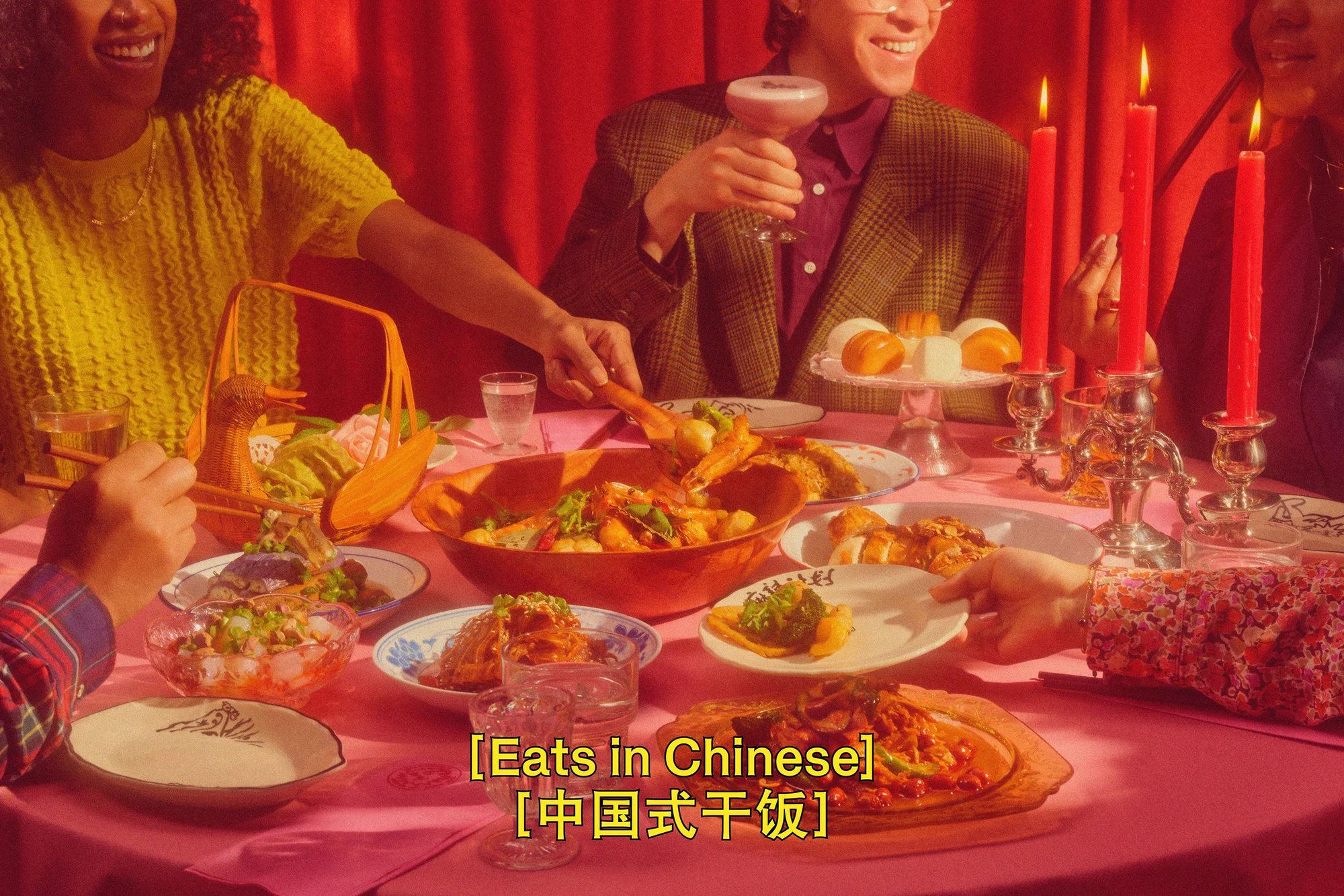 Several people sit at a table laden with food, drinks, candles and decorative objects in an indoor setting with red curtains.