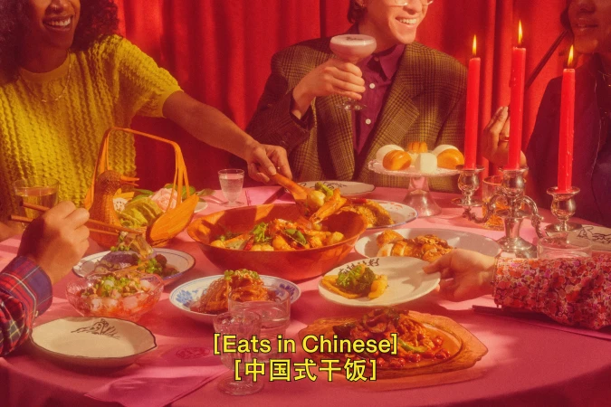 Several people sit at a table laden with food, drinks, candles and decorative objects in an indoor setting with red curtains.